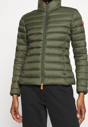 Personne portant une veste matelassée ajustée vert olive avec une tirette de fermeture éclair orange et un pantalon noir, vue des épaules aux hanches.