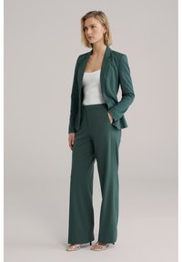 Žena odjevena u tamnozeleno krojeno odijelo s širokim hlačama, bijelu majicu bez rukava i bijele sandale s remenčićima, stoji s jednom rukom u džepu.