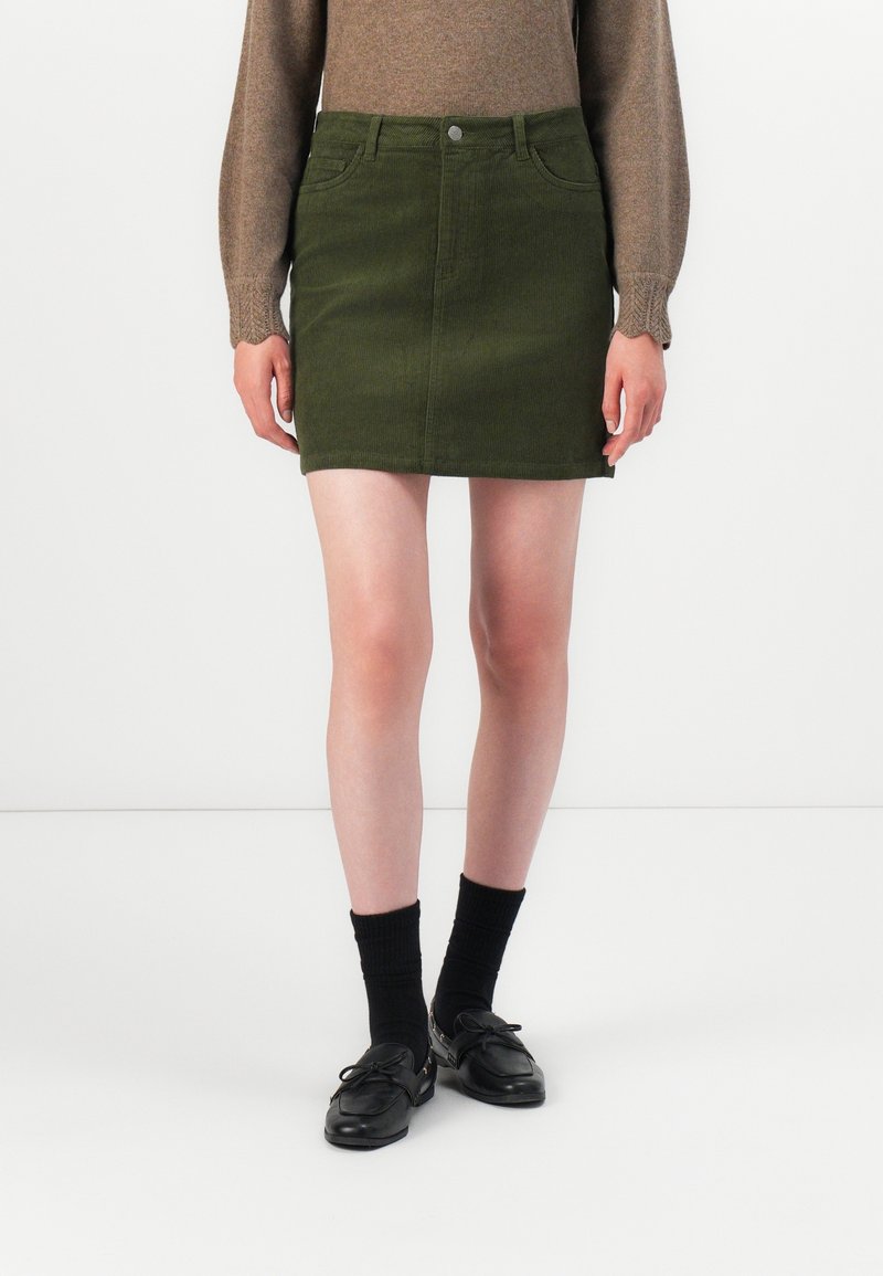 Personne portant une mini-jupe en velours côtelé vert olive, un haut marron à manches longues, des chaussettes noires et des mocassins noirs, debout sur un sol blanc.