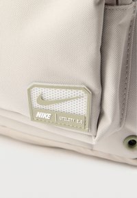 Γωνία από μπεζ υφασμάτινη τσάντα Nike Utility 2.0 που δείχνει υφή υλικού και ετικέτα από καουτσούκ με το λογότυπο Swoosh.