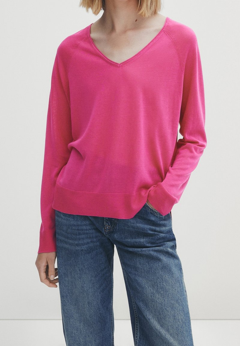 Pull col en V fuchsia à manches longues, poignets et ourlet côtelés, texture en maille légère, associé à un jean bleu à jambes larges.