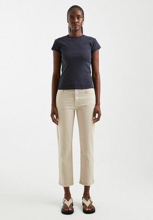 Kvinde stående med fronten fremad iført en navy t-shirt, beige korte bukser og beige sandaler mod en ensfarvet lys baggrund.