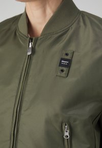 Olivově zelená bomber bunda se zipem, žebrovaným límcem a značkovou etiketu. Obsahuje boční kapsy se kovovými zipy, hladkou texturu látky.