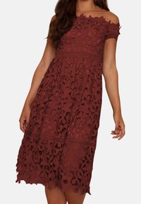 Punainen olan paljastava mekko, jossa on kerroksittain kukkakuvioista pitsiä, istuva yläosa, teksturoitu kangas ja kerroksellinen helma.