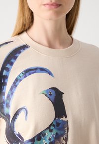 Sudadera de algodón beige que presenta un gráfico de un pájaro colorido en tonos azules y verdes, con un escote redondeado y detalles acanalados.