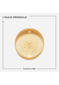 Goldener Öltropfen mit sichtbaren Bläschen, zentriert auf weißem Hintergrund, beschriftet mit "L'HUILE ORIGINALE" und "ELIXIR ULTIME" in schwarzem Text.