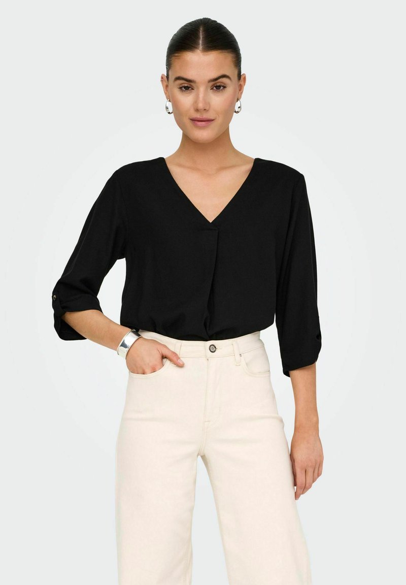 Schwarze V-Ausschnitt-Bluse mit drei Viertelärmeln und Knopfdetails, kombiniert mit hellbeigen Hosen. Glatte Textur, lockere Passform.