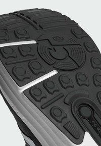 Suela de goma negra con superficie texturizada, que presenta patrones circulares y lineales, y una franja blanca a lo largo del costado. Incluye un detalle notable del logotipo.