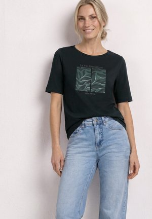 Smilende kvinde med blondt hår iført sort grafisk t-shirt og lyseblå jeans, stående mod en ensfarvet lys væg.