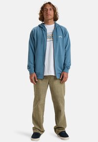 Sweat à capuche zippé bleu clair avec poches avant, porté sur un t-shirt graphique blanc. Associé à un pantalon en velours côtelé beige et des chaussures foncées.