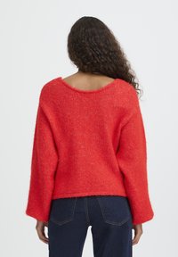Maglione rosso lavorato a maglia con scollo ampio e maniche lunghe. Texture morbida, vestibilità ampia e leggermente corto nella parte posteriore, abbinato a jeans scuri.
