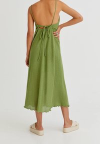 Kobieta ubrana w zieloną, teksturowaną sukienkę midi z cienkimi ramiączkami związanymi z tyłu, w połączeniu z beżowymi sandałami na platformie, stojąca na białej podłodze.