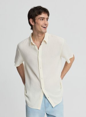 Giovane uomo sorridente, indossa una camicia a maniche corte color avorio con trama e bottoni e jeans azzurri chiari, in piedi con le mani dietro la schiena.