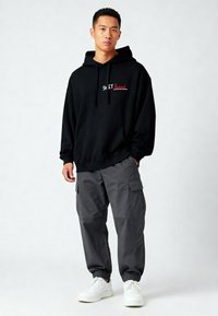 Felpa nera con testo bianco e rosso sul petto, abbinata a pantaloni cargo grigi e sneakers bianche. Design casual e veste ampia.