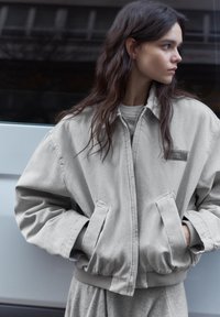 Junge Frau mit langen dunklen Haaren, trägt eine übergroße hellgraue Jacke, Hände in den Taschen, schaut nach rechts vor einem dunklen Hintergrund.