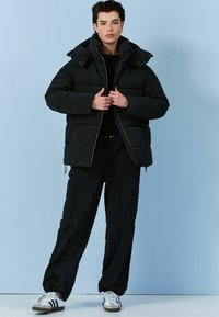 Melna pūkains mētelis ar augstu apkakli un priekšējo rāv�ējslēdzi, apvienots ar brīviem melniem kravas biksēm un baltām kedām. Visā virsmā ir gluda tekstūra.