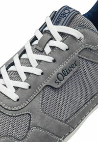 Baskets grises avec un tissu texturé et des accents en cuir, lacets blancs et une étiquette logo bleue. Présente une couture contrastée le long des bords.