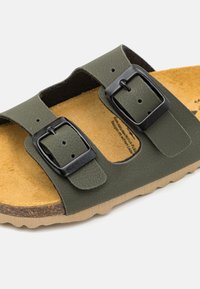 Sandale synthétique verte avec deux boucles noires ajustables, un semelle intérieure texturée beige et une semelle en caoutchouc marron clair avec un motif ondulé.