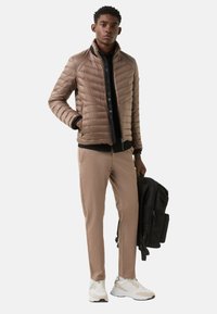Steppjacke in Hellbraun mit horizontalen Steppnähten, schwarzes Unterhemd, beige Hose und weiße Turnschuhe. Trägt einen schwarzen Rucksack.