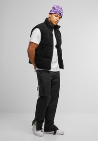 Svart fleecestoff vest over en hvit t-skjorte, kombinert med svarte cargo-bukser med sidelommer. Lilla tie-dye lue og hvite sneakers.