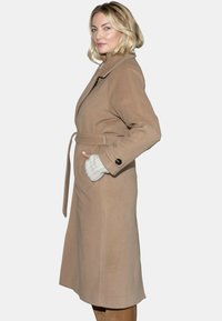 Długi, beżowy płaszcz wełniany z pasem w talii, dużym kołnierzem i bocznymi kieszeniami. Udekorowany czarnym guzikiem na mankiecie. Gładka textura.