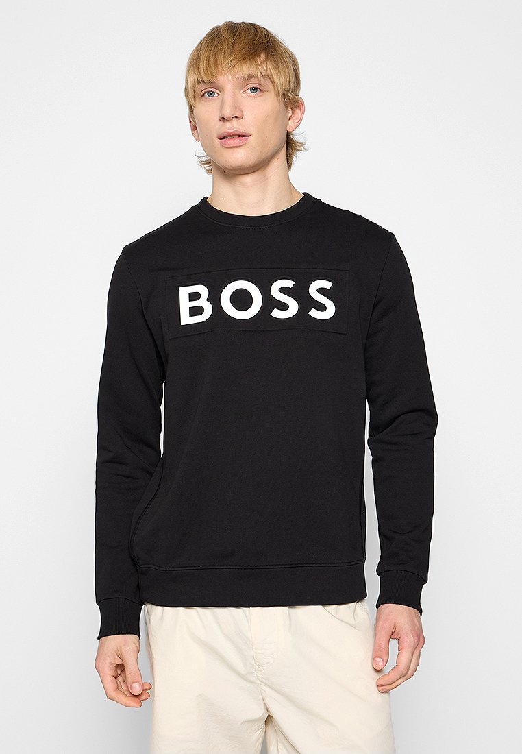 Un giovane uomo indossa una felpa nera con la scritta "BOSS" in lettere bianche e pantaloni beige chiaro, in piedi contro uno sfondo chiaro e uniforme.