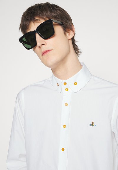 White shirt with a pointed collar, featuring orange buttons and a small embroidered design. Black angular sunglasses with green lenses.