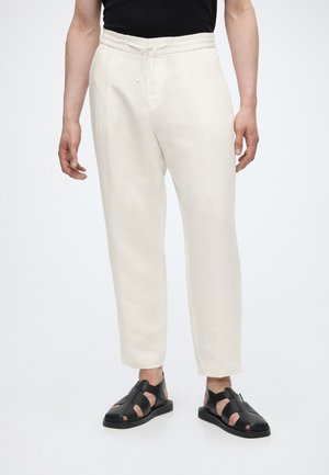 Homme portant un pantalon à cordon écru et des sandales en cuir noires, debout devant un fond clair uni.