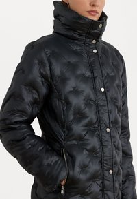 Black puffer jacket with a high collar, quilted pattern, silver snap buttons, and a zippered pocket on the left side. Smooth texture.