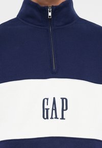 Námořnický zipový svetr s bílým vodorovným pruhem; má výšivku loga "GAP" v námořnické barvě, hladkou texturu a vysoký design límce.