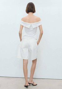 Witte off-the-shoulder top met een strikdetail, gecombineerd met witte shorts. Het model draagt zwarte hakken. Glad materiaal, minimalistisch ontwerp.