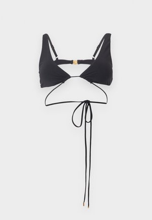 Vrouw met nat haar die een zwarte tweedelige bikini met omwikkelde bandjes draagt, staande tegen een egale lichtgrijze achtergrond.