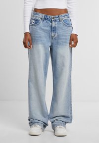 Světle modré široké denimové džíny s vybledlým efektem, s předními kapsami a značkovou nášivkou na pasu. Nosené s bílými teniskami.