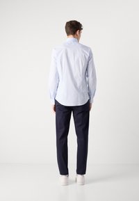 Hombre de pie de espaldas, vistiendo una camisa de manga larga azul clara, pantalones oscuros de color marino y zapatillas blancas, contra un fondo blanco.