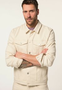 Chaqueta de denim beige con dos bolsillos en el pecho, cierres de botón y mangas dobladas. Presenta un cuello clásico y un diseño estructurado.