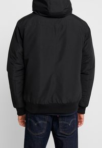 Sort hættepufferjakke med en glat yderside, elastiske manchetter og kant. Har et simpelt design og et let struktureret stof.