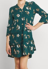 Tealfärgad blommig klänning med trekvartsärmar, v-ringning, volangdetaljer och avslappnad passform. Har lätta blommor och fjärilstryck.