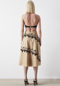 Beige A-lijn rok met zwart abstract patroon langs de zoom, gemaakt van gladde stof. Draag het met een zwart crop top en zwarte haksandalen.