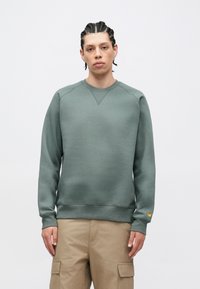 Žalias džemperis su apvalia iškirpte, raglano rankovėmis, tekstūruota medžiaga, ribotomis rankogaliais ir apačia, kairėje rankovėje mažas auksinis logotipas.