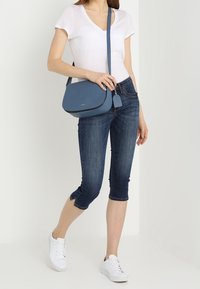 Blå axelväska i texturerat material med en rundad form och dragkedja. Bärs med vit t-shirt och mörka cropped jeans. Vita sneakers.