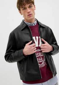 Schwarze Lederjacke mit Kragen, Reißverschlusstaschen und lässiger Passform, getragen über einem bordeauxfarbenen Sweatshirt mit weißem Text.