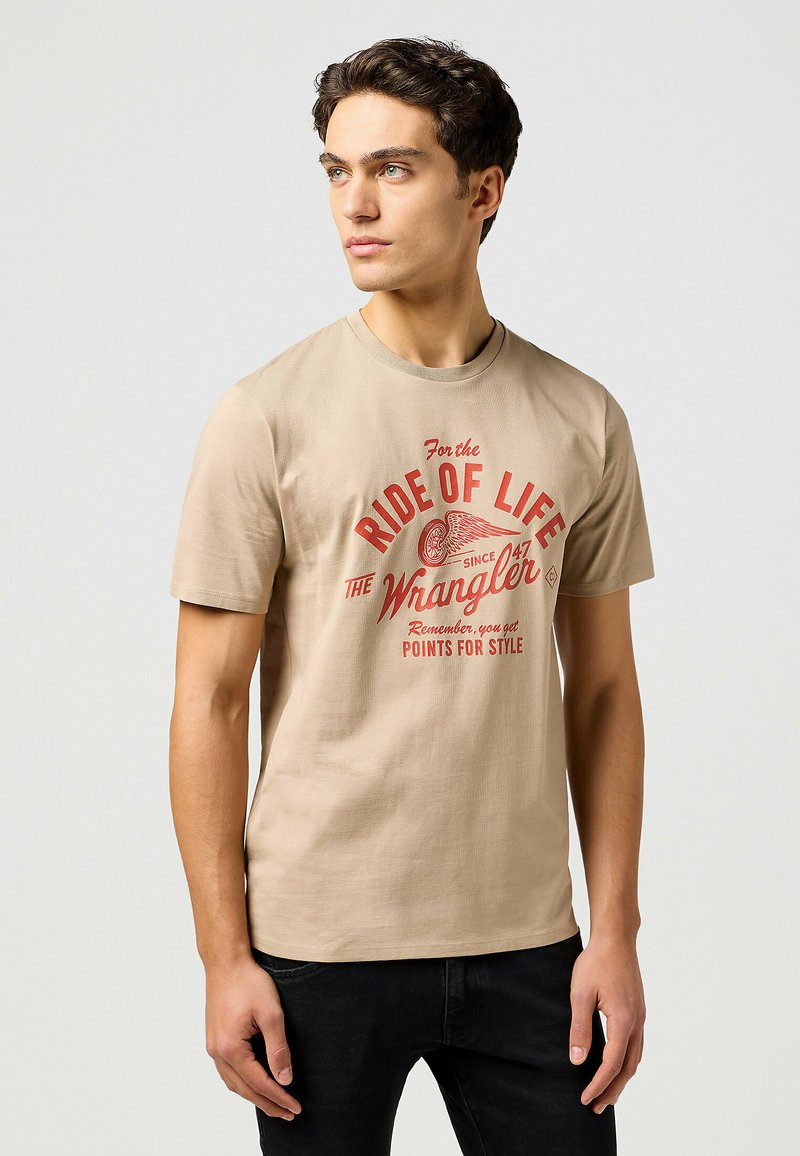 Vaahto puuvillasta valmistettu beige t-paita, jossa on pyöreä pääntie. Etupuolella on punaisella tekstillä grafiikka, jossa lukee "For the Ride of Life" ja "Wrangler."
