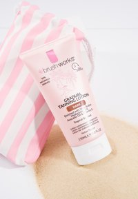 Tube de lotion bronzante progressive étiquetée "sombre" avec un design de flamant rose et de palmier, placée sur du sable à côté d'un sac à cordon rayé rose et blanc.