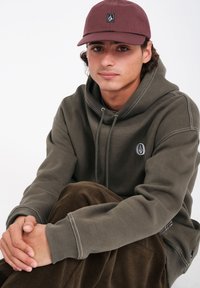 Olijfgroene hoodie met witte stiksels, met een klein logopatch; bruine corduroy broek. Model zit en draagt een kastanjebruine pet.