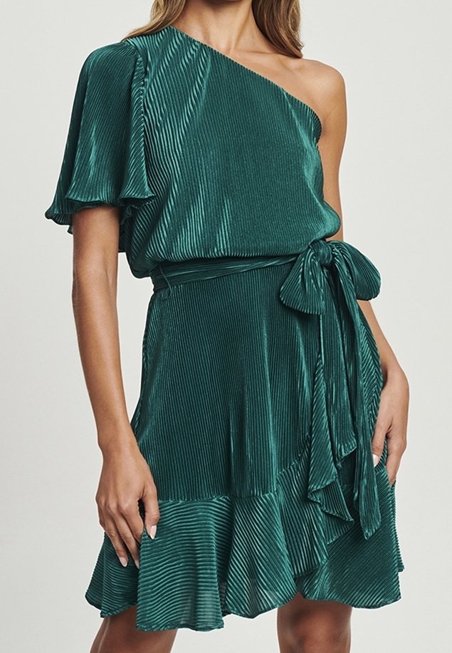Femme portant une robe plissée turquoise à une épaule avec un ourlet à volants et un nœud à la taille, debout devant un fond uni.