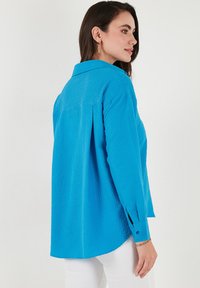 Camicia blu con texture, maniche lunghe, cucitura a yoke sul retro e polsini con bottoni. Presenta una vestibilità rilassata e orlo arrotondato.
