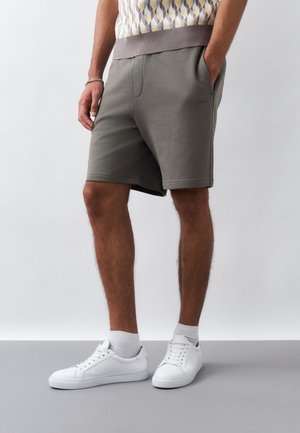 Άνδρας που φοράει γκρι καθημερινές βερμούδες, λευκές κάλτσες χαμηλές και λευκά αθλητικά παπούτσια με κορδόνια, στέκεται με τα χέρια στις τσέπες πάνω σε γκρι πάτωμα.