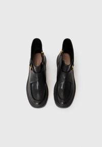 Pantofi negri din piele, fără siret, cu vârf rotund, având un design încrucișat, accente din hardware auriu și o talpă groasă neagră.