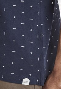 Camisa de manga corta azul marino con un tejido texturizado que presenta formas geométricas blancas y la palabra "VIERNES" en un patrón repetido. Etiqueta mostrada.