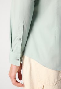 Tricou verde deschis cu mânecă lungă, având o textură netedă, mansete aberate și o margine ușor curbată, prezentat cu o piesă vestimentară alb-off dedesubt.